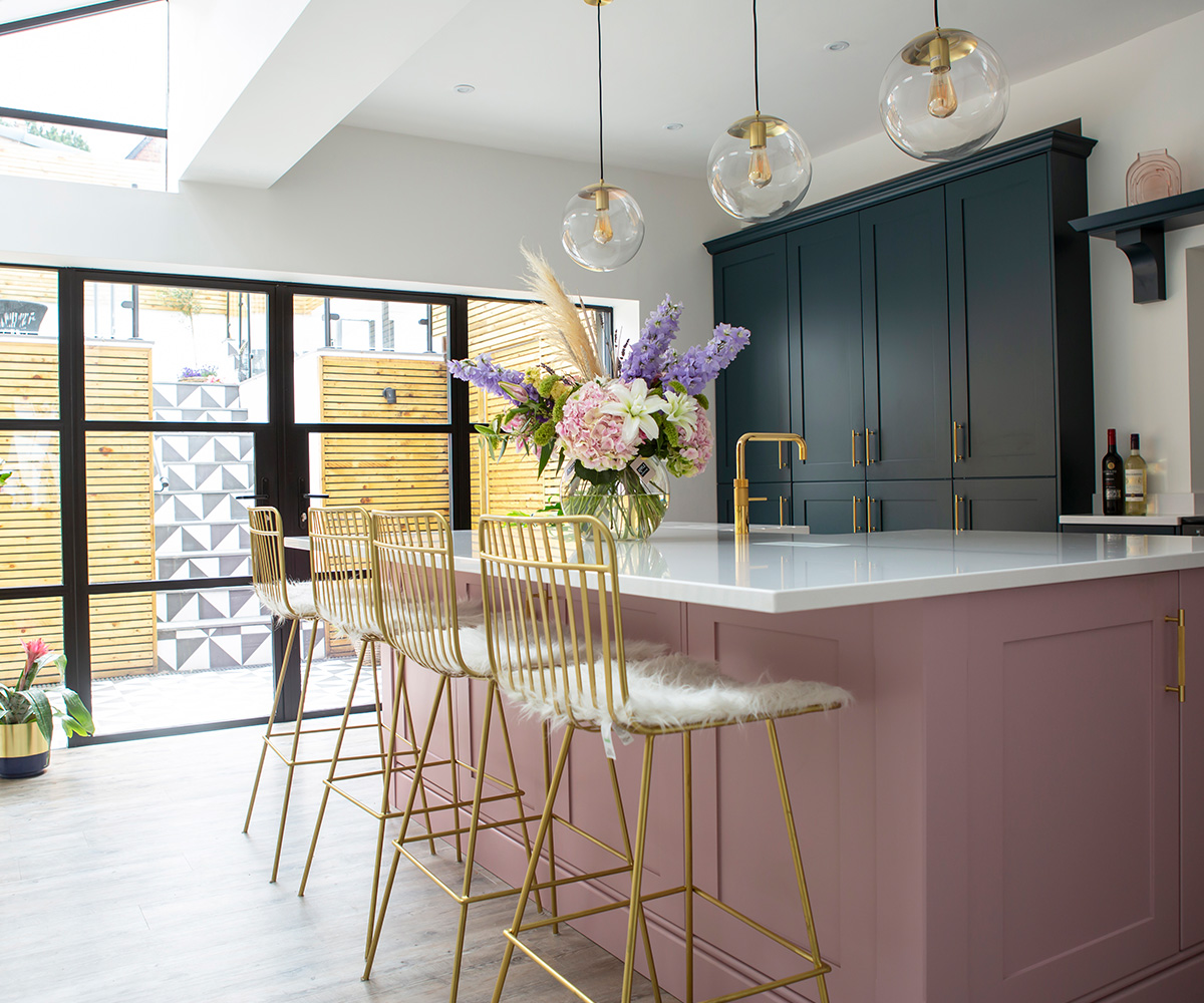 A real customer kitchen featuring a light pink kitchen island with gold bar stools. in the back there are dark blue kitchen units.